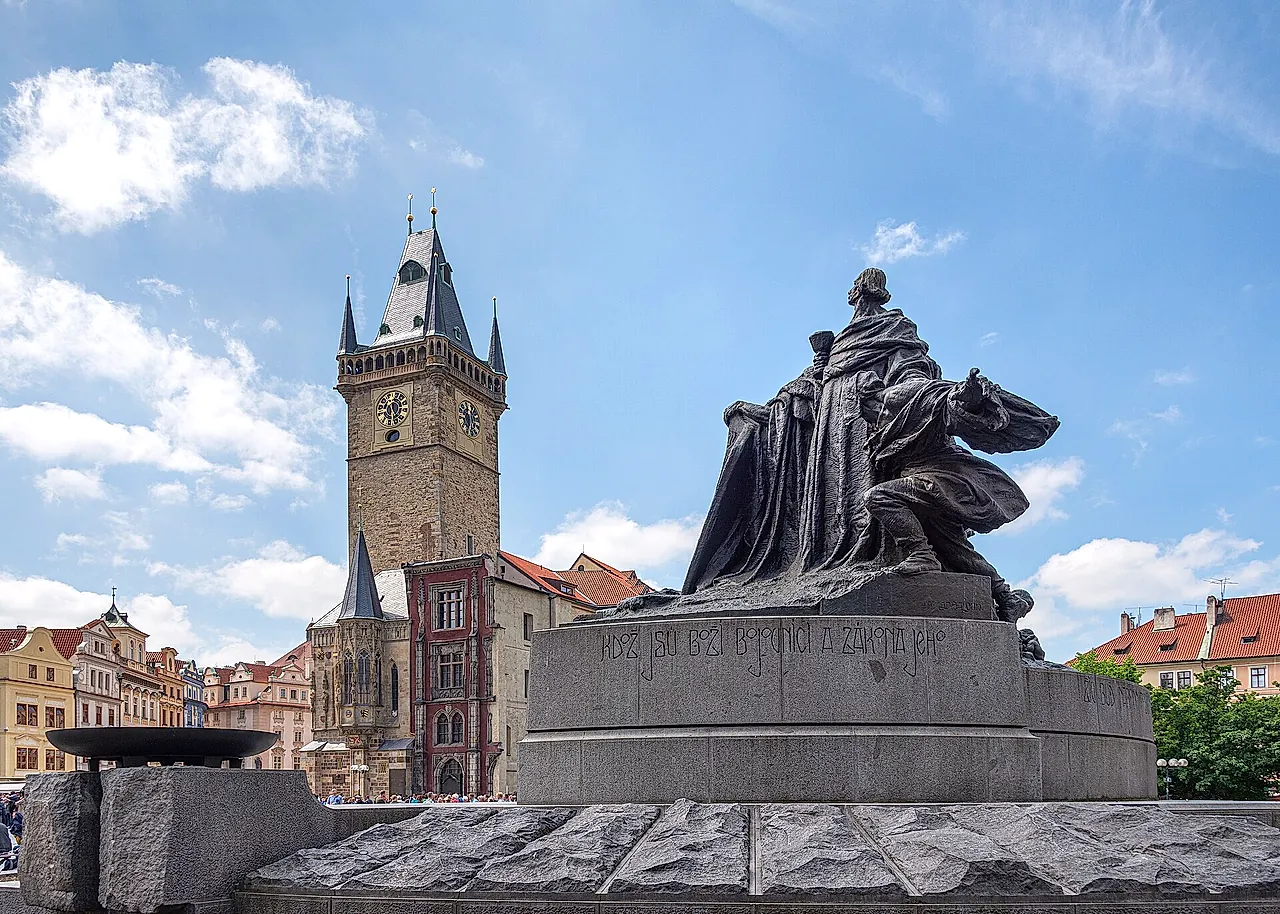 The Jan Hus monument in Old Town Square, a bronze figure on a granite plinth, with the Old Town Hall tower rising behind it under a blue sky.