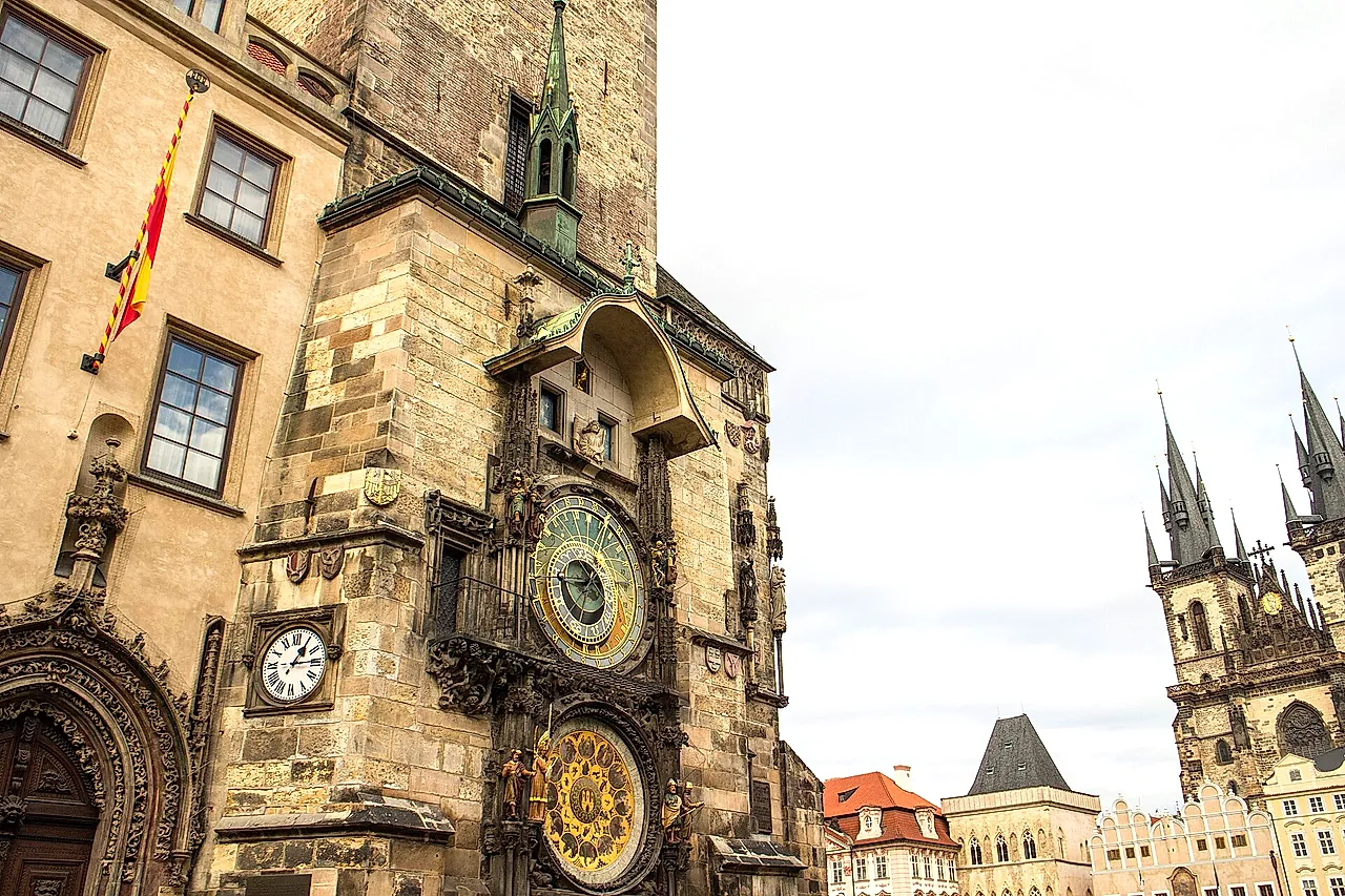 Astronomical clock on the Old Town Hall tower, seen from the side, with its blue astronomical dial and calendar dial below, and the spires of Týn Church beyond.