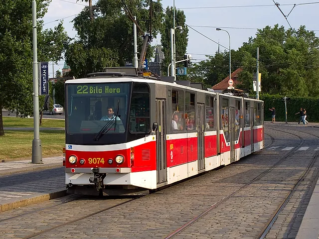A red-and-white tram on line 22 with its destination board reading Bílá Hora, running on cobbled tracks beside a grassy strip and tall trees.
