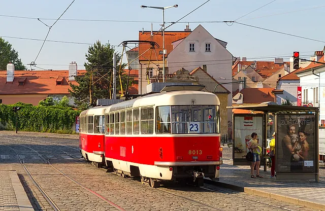 Red-and-cream tram 8013 on line 23 waiting at a stop, with a glass shelter on the right and orange-roofed houses behind under a pale sky.