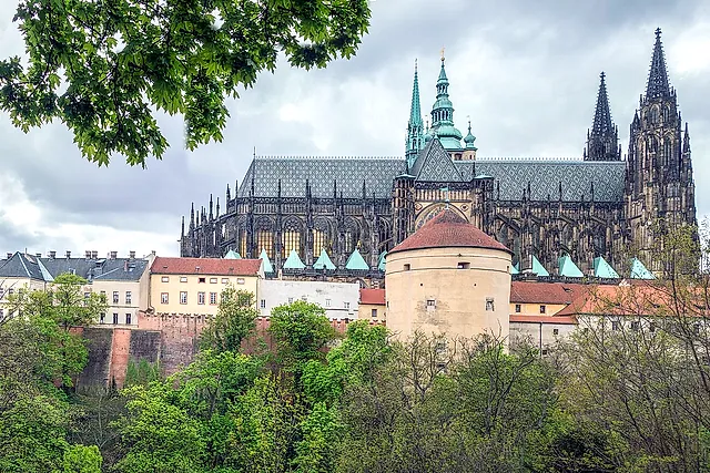 St Vitus Cathedral from the north side, with Gothic flying buttresses and two spires above a round stone tower with a red roof, under a cloudy sky.