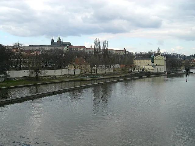 The Vltava flowing past an embankment wall and riverside trees, with Prague Castle and its cathedral spires on the hill beyond under a cloudy sky.