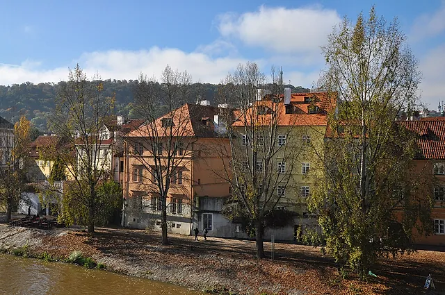 Riverside houses in orange and cream along the Vltava at Kampa, with leafless trees in front and wooded hills rising behind under a blue sky with clouds.