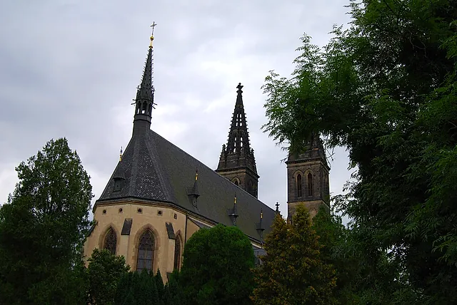 Gothic church with a steep grey roof and two pointed spires rising above green trees and a lower stone chapel with arched windows, under a cloudy sky.