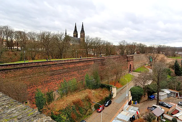 Red brick fortress wall of Vyšehrad curving toward a gate, with the twin spires of the basilica above the trees and parked cars on the road below.