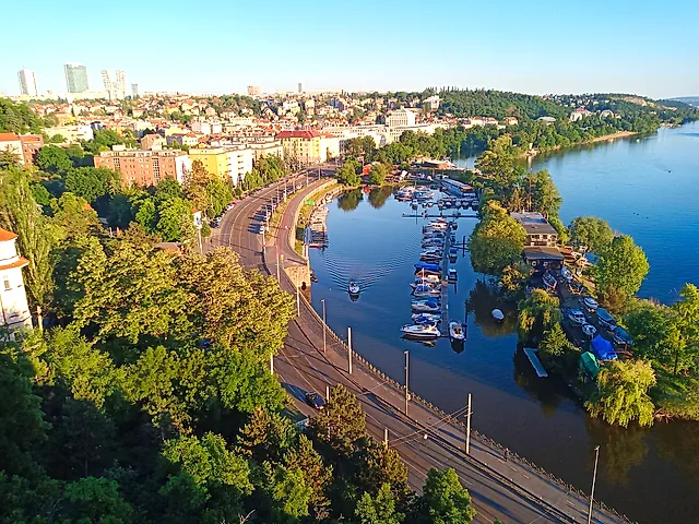 Aerial view of a Vltava marina with moored boats, a riverside road lined with trees, and city blocks and wooded hills beyond under a clear blue sky.