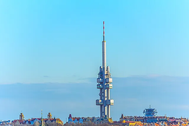 The Žižkov Television Tower seen from a distance against a pale blue sky, its concrete pods and tall mast rising above the rooftops of the city.