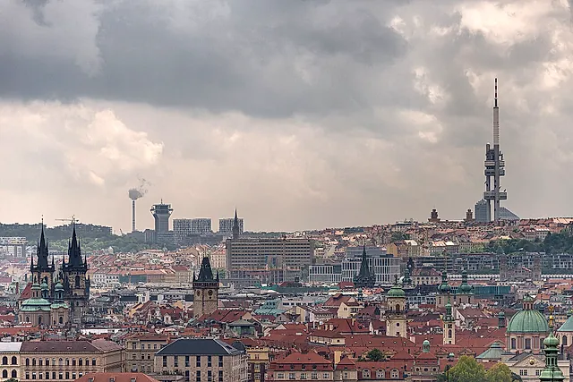 The Žižkov Television Tower on the right of a view across Prague's red roofs, with the twin spires of Týn Church at left under a heavy grey sky.
