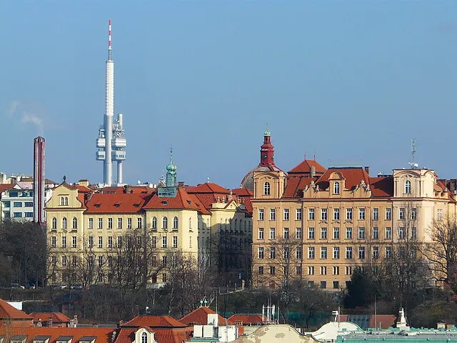 View from Vyšehrad over Prague's rooftops, with the Žižkov Television Tower rising on the left above pale ochre buildings under a clear blue sky.